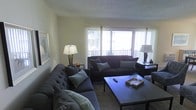 This single-floor space features a comfortable and thoughtfully arranged living room as its central gathering area. The room is anchored by two generously sized dark upholstered sofas, one of which is a tufted design, both dressed with coordinating throw pillows in varying shades of grey and blue. A rectangular wooden coffee table sits at the center, styled with a decorative tray, glassware, and a remote control, adding a functional yet polished touch.

Large windows with sheer white curtains span much of one wall, flooding the room with abundant natural light and offering a view of an outdoor balcony railing and surrounding buildings beyond. The bright, airy quality of the space is enhanced by the pale yellow-toned walls and a textured ceiling overhead.

Warm ambient lighting is provided by two table lamps — one positioned beside each seating area — contributing to an inviting atmosphere during evening hours. A leopard-print area rug grounds the seating arrangement and adds a subtle decorative accent. Wall art and a dark side table with additional décor complete the space, giving it a lived-in, welcoming character.