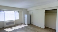 This single-floor space presents a clean, open layout with a minimalist aesthetic. The interior is finished with light-colored walls and wall-to-wall carpeting, contributing to a neutral and functional atmosphere. A window fitted with horizontal blinds allows natural light to filter into the room, casting soft sunlight across the floor and enhancing the sense of openness within the space.

A full-sized white refrigerator is positioned along one wall, indicating a combined living and kitchen area typical of a studio-style layout. Adjacent to the window, a wall-mounted HVAC unit provides climate control directly within the main living area. Structural details include a recessed ceiling border that frames the room, adding subtle architectural definition to the otherwise understated interior.

The space is currently unfurnished, allowing for flexible furniture arrangement and personalization. The door visible along the far wall suggests access to an additional area or closet, adding functional storage potential. Overall, the space offers a straightforward, light-filled environment well-suited for practical, comfortable living.