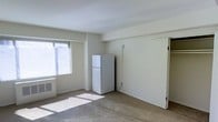This single-floor space presents a clean, open layout with a minimalist aesthetic. The interior is finished with light-colored walls and wall-to-wall carpeting, contributing to a neutral and functional atmosphere. A window fitted with horizontal blinds allows natural light to filter into the room, casting soft sunlight across the floor and enhancing the sense of openness within the space.

A full-sized white refrigerator is positioned along one wall, indicating a combined living and kitchen area typical of a studio-style layout. Adjacent to the window, a wall-mounted HVAC unit provides climate control directly within the main living area. Structural details include a recessed ceiling border that frames the room, adding subtle architectural definition to the otherwise understated interior.

The space is currently unfurnished, allowing for flexible furniture arrangement and personalization. The door visible along the far wall suggests access to an additional area or closet, adding functional storage potential. Overall, the space offers a straightforward, light-filled environment well-suited for practical, comfortable living.