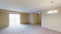 This 1-bedroom, 1-bathroom residence features a thoughtfully designed G Style floor plan across a single level. The open living area is a central highlight, showcasing plush wall-to-wall carpeting and warm neutral-toned walls that create a welcoming atmosphere. A large sliding glass door floods the space with natural light while providing direct access to the outdoors, enhancing the sense of openness throughout. A ceiling-mounted light fixture provides additional illumination, and a built-in HVAC vent ensures year-round climate comfort. Adjacent to the main living area, a defined transition zone features parquet-style hard flooring, adding a subtle contrast in texture and material that helps delineate functional areas within the open layout. Clean white trim and baseboards frame the rooms with a crisp, finished look. The overall design is practical and inviting, offering a straightforward layout well-suited for comfortable everyday living. The neutral palette and open spatial flow provide a versatile canvas that accommodates a variety of furnishing styles and personal preferences.