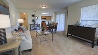 This single-floor residence offers a thoughtfully designed open-concept layout that seamlessly connects the living, dining, and kitchen areas, creating a spacious and functional environment. The living area is anchored by plush wall-to-wall carpeting and features a comfortable seating arrangement centered around a glass-top coffee table with wrought iron legs, complemented by decorative wall mirrors and tasteful accent furniture. Natural light fills the space through large sliding glass doors dressed with vertical blinds, lending the interior a bright and welcoming ambiance. Adjacent to the living area, the dining space accommodates a dark wood table with upholstered chairs, ideal for everyday meals or entertaining. A skylight above the kitchen passthrough enhances the natural illumination throughout the central living zone. The kitchen is situated just beyond, featuring warm wood cabinetry, black appliances, and a practical layout. Neutral-toned walls and light ceiling fixtures maintain a cohesive and understated aesthetic throughout. A hallway visible from the living area leads to additional private spaces, completing this well-balanced and move-in-ready home.
