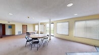 Located at 1749 Lynx Ave in Webster City, Iowa, this single-floor space is designed to accommodate gatherings and community activities. The main area is an open, carpeted room with soft yellow walls and a textured ceiling, illuminated by recessed lighting and an abundance of natural light streaming through multiple large windows fitted with horizontal blinds. Structural columns are thoughtfully integrated into the layout, and the room comfortably accommodates multiple round tables with folding chairs, making it well-suited for meetings, events, or social gatherings. A glass door provides direct access to the outdoors, offering views of the surrounding landscape. The space also features a dedicated kitchen, finished with rich dark-wood cabinetry, granite-style countertops, a double stainless steel sink, and durable tile flooring. Ample upper and lower cabinetry provides generous storage, and a large window keeps the kitchen bright and pleasant. Safety features including a fire extinguisher and exit signage are present throughout. The overall atmosphere is clean, functional, and welcoming, making this a practical and versatile space for a variety of community or organizational uses.