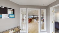 This generously sized single-story community space is thoughtfully designed to serve as a social hub for residents. The layout flows seamlessly from a welcoming entry foyer — complete with an informational display board and a small side table for literature — into a bright, open main lounge area. The lounge is anchored by a built-in fireplace framed by a flat-screen television and flanked by French doors that allow natural light to fill the room. Light hardwood-style flooring runs throughout the entire space, creating a cohesive and clean aesthetic against the neutral gray walls.

The main gathering area accommodates a comfortable seating arrangement with upholstered sofas and accent chairs, while a separate dining zone offers multiple round tables with decorative wrought-iron chairs, perfect for casual meals or group activities. Recessed lighting throughout ensures the space is well-lit at all times.

The fully equipped kitchen features warm wood cabinetry, stainless steel appliances including a refrigerator, microwave, and range, as well as a peninsula with bar seating for two — ideal for hosting events or community meals. Horizontal transom windows in the kitchen bring in soft natural light.

Additional areas include an open reception or service counter and a dedicated lounge corner, making this a versatile and functional community amenity space.