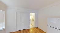 This single-floor space is designed with a clean, minimalist aesthetic, highlighted by crisp white walls and smooth white ceilings that create a bright, airy atmosphere. The main living area features warm-toned hardwood flooring that adds a sense of natural warmth and continuity throughout the space. A set of classic six-panel double doors provides generous closet storage, finished in white to blend seamlessly with the surrounding walls.

An open doorway connects the main living area directly to the kitchen, which is efficiently laid out and well-lit. The kitchen is outfitted with white upper and lower cabinetry, offering ample storage, and includes a built-in sink with a countertop workspace. Light-toned tile flooring in the kitchen contrasts subtly with the hardwood in the main area, clearly defining the two zones. The kitchen's warm overhead lighting contributes to an inviting atmosphere.

Overall, the space maintains a cohesive, neutral color palette throughout, emphasizing functionality and simplicity. The layout is practical, with thoughtful transitions between areas making the space feel organized and easy to navigate.