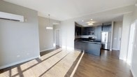 This single-floor residence showcases a thoughtfully designed open-concept layout that seamlessly blends the living and kitchen areas into one cohesive, light-filled space. Warm hardwood flooring runs throughout, adding a natural contrast to the cool, contemporary aesthetic of the interior.

The kitchen is a standout feature, outfitted with dark charcoal cabinetry, dark stone countertops, and a full suite of stainless steel appliances, including a French door refrigerator, stainless range, and built-in microwave. A kitchen island provides additional workspace and visual separation between the cooking and living areas. Recessed lighting and a flush ceiling fixture illuminate the kitchen zone effectively.

The adjacent living area is bright and airy, with neutral gray walls and abundant natural light streaming in, casting long, warm rays across the hardwood floors. A stylish rectangular pendant light anchors the dining area, adding a modern touch to the open floor plan.

Clean architectural lines, white baseboards, and a neutral color palette throughout create a calm, sophisticated ambiance. The space flows naturally, making it well-suited for both everyday living and entertaining.