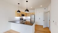Located on West Broad Street in Richmond, Virginia, this well-appointed one-bedroom, one-bathroom residence offers a thoughtfully designed single-floor layout with modern finishes throughout. Light hardwood flooring flows continuously across the open-concept kitchen and living area, creating a cohesive and airy feel.

The kitchen is a standout feature, anchored by a peninsula with rich dark granite countertops and an undermount sink, illuminated by two striking matte black dome pendant lights. Light maple upper cabinetry contrasts cleanly with white lower cabinets, while a white subway tile backsplash and a full suite of stainless steel appliances — including a refrigerator, range, microwave, and dishwasher — complete the space.

The bedroom offers a comfortable footprint with light-toned hardwood floors, a ceiling fan, and a large window framing open city views, filling the room with natural light. A hallway connects the main living areas and provides access to the full bathroom, which features a soaking tub with a vertically laid blue tile surround, a granite vanity, and tile flooring. A dedicated in-unit laundry closet with a stacked washer and dryer adds convenience. Clean white walls and recessed lighting throughout give the entire residence a bright, contemporary character.