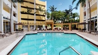 This outdoor pool area exudes a tropical resort atmosphere, featuring a generously sized, crystal-clear swimming pool with calm turquoise water that reflects the surrounding greenery and open sky. The pool is complemented by a shallow entry section, making it accessible and inviting. Surrounding the pool is a spacious tiled deck furnished with lounge chairs and seating areas, ideal for sunbathing or unwinding in the open air. Tall, mature palm trees are positioned throughout the space, providing natural shade and enhancing the tropical ambiance. A covered pergola-style structure in the background serves as a shaded gathering area, adding architectural interest and a comfortable retreat from direct sunlight. The warm yellow facade of the adjacent multi-story building anchors the space visually, while lush greenery along the perimeter creates a sense of seclusion. Potted plants and carefully maintained landscaping contribute to the polished, resort-like aesthetic. The overall atmosphere is bright, open, and relaxing, well-suited for leisure and recreation in a warm, sun-drenched environment.