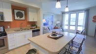 This single-floor residence showcases an open-concept layout that connects the kitchen and living areas into one cohesive, functional space. The kitchen is outfitted with white cabinetry, a stainless steel dishwasher, and an undermount sink set against a warm terracotta accent wall that adds a pop of color and visual interest. A spacious granite-topped island anchors the kitchen, providing additional prep and casual dining space, illuminated by two stylish pendant lights hanging overhead. The living area flows naturally from the kitchen, creating an inviting atmosphere ideal for both relaxing and entertaining. A comfortable sofa, decorative artwork, and coordinating accent pieces furnish the living space, while large windows allow abundant natural light to fill the room and provide views of the outdoors. Light hardwood-style flooring runs throughout, complementing the neutral color palette and tying the spaces together. The overall ambiance is modern yet welcoming, combining practical design elements with thoughtful aesthetic touches that make the space feel both functional and livable.