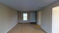 This two-story residence offers a straightforward and functional layout across two floors. The main level greets visitors with a front entry door finished in a clean grey tone, providing a welcoming point of arrival. The living area is generously proportioned, featuring wall-to-wall carpeting that adds warmth underfoot. Neutral grey-toned walls create a calm, versatile backdrop throughout, complemented by crisp white baseboards that frame each room cleanly. A window on the main level allows natural light to filter into the living space, offering a view of the surrounding greenery outside. Ceiling vents are integrated discreetly into the layout, supporting climate control throughout the home. A staircase positioned near the entry provides access to the upper floor, allowing for a clear separation between the living areas on each level. An adjacent doorway leads to additional rooms, suggesting further functional spaces beyond the main living area. Overall, the residence presents a clean, move-in ready aesthetic with a neutral palette well-suited for a variety of interior styles.