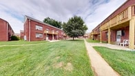 Located at 7003 Dunmanway in Dundalk, Maryland, this multi-building brick residential complex showcases a timeless architectural style characterized by sturdy red brick exteriors, neutral rooflines, and wooden balconies that add warmth and character to the façade. The buildings are arranged to frame a generous central courtyard, creating an open, community-oriented atmosphere that encourages outdoor activity and neighborly interaction.

The shared outdoor grounds are a defining feature of this property. Expansive, well-maintained lawns stretch between the buildings, dotted with mature shade trees that provide natural coverage and a sense of established greenery. Concrete walkways wind throughout the complex, offering easy pedestrian access to all areas of the grounds. Street-style lamp posts are positioned along the pathways, ensuring safe and well-lit navigation after dark.

Each building features individual unit entrances at ground level alongside upper-level balconies with wooden railings, providing residents with private outdoor space. On-site parking is readily available, adding to the practical convenience of the property. A prominent brick chimney tower serves as a distinctive architectural landmark visible throughout the complex. The overall ambiance is that of a cohesive, close-knit residential community with functional amenities and ample open space.