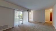 This single-story space is designed with a straightforward, functional layout that connects several distinct living areas. The main living room is generously sized, featuring wall-to-wall neutral-toned carpeting and a full-width sliding glass door dressed with vertical blinds. The door allows abundant natural light to fill the room and provides direct views of outdoor greenery. A ceiling fan is visible in the adjacent area, which flows seamlessly from the main living space, creating an open, connected feel throughout.

The bedroom is a well-proportioned room with the same neutral carpeting, white walls, and a window fitted with vertical blinds that bring in natural light. A door leads to a connecting hallway, offering convenient access to the rest of the unit.

The kitchen features warm wood-toned cabinetry both above and below the countertops, a stainless steel single-basin sink, and light countertop surfaces with ample workspace. Upper cabinets provide practical storage.

The bathroom is tiled throughout, with white square tiles lining the walls around the bathtub and shower area. It includes a standard tub, toilet, vanity with a wood cabinet base, and a mirrored medicine cabinet above the sink. The overall space maintains a clean, practical aesthetic throughout.