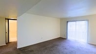 This single-floor space presents a clean, functional layout designed for comfortable everyday living. The main living area is generously proportioned, featuring wall-to-wall plush carpeting in a neutral tone that adds warmth underfoot. A notable architectural detail is the angled ceiling line along one wall, adding visual interest to the otherwise straightforward design. A full-height sliding glass door fitted with vertical blinds draws in abundant natural light and provides access to an outdoor area, brightening the interior considerably.

The neutral white walls throughout create a bright, versatile backdrop that adapts easily to various furnishing styles. The transition from the living area leads into a carpeted hallway, where louvered bifold doors — framed in contrasting dark trim — suggest convenient storage or utility closet access. The consistent use of neutral tones across walls, ceilings, and flooring gives the space a cohesive, understated aesthetic. Multiple doorways visible along the hallway indicate a well-connected floor plan, with rooms flowing logically from one to the next. Overall, the space offers a straightforward and adaptable environment suitable for a range of uses.