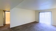 This single-floor space presents a clean, functional layout designed for comfortable everyday living. The main living area is generously proportioned, featuring wall-to-wall plush carpeting in a neutral tone that adds warmth underfoot. A notable architectural detail is the angled ceiling line along one wall, adding visual interest to the otherwise straightforward design. A full-height sliding glass door fitted with vertical blinds draws in abundant natural light and provides access to an outdoor area, brightening the interior considerably.

The neutral white walls throughout create a bright, versatile backdrop that adapts easily to various furnishing styles. The transition from the living area leads into a carpeted hallway, where louvered bifold doors — framed in contrasting dark trim — suggest convenient storage or utility closet access. The consistent use of neutral tones across walls, ceilings, and flooring gives the space a cohesive, understated aesthetic. Multiple doorways visible along the hallway indicate a well-connected floor plan, with rooms flowing logically from one to the next. Overall, the space offers a straightforward and adaptable environment suitable for a range of uses.