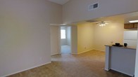 This single-floor residential space is defined by its impressive vaulted ceilings that dramatically elevate the main living area, creating a sense of openness and volume rarely found in similarly sized homes. The living room is generously proportioned, finished with wall-to-wall plush carpeting in a neutral tone that extends seamlessly throughout the space, providing a warm and cohesive feel underfoot.

A ceiling fan with a light fixture is positioned in the adjoining area, partially separated by a structural beam detail that subtly delineates the living and dining zones while maintaining an open-concept flow. HVAC vents are integrated into both the ceiling and upper wall, ensuring efficient climate control throughout.

An open doorway leads to an additional carpeted room, likely a bedroom, which benefits from natural light streaming through a window dressed with vertical blinds. The neutral, light-toned walls throughout create a clean, versatile backdrop that adapts well to a variety of interior styles.

A partial view of what appears to be a countertop or bar area is visible along one edge, suggesting a kitchen or breakfast bar nearby. The overall ambiance is calm and functional, offering a straightforward yet comfortable layout well-suited for everyday living.
