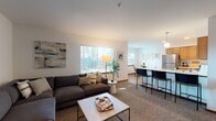 This single-floor residence offers a thoughtfully designed layout that balances comfort and functionality. The open-concept living area is anchored by large windows that flood the space with natural light, offering views of the surrounding greenery. The living room provides generous seating space and connects seamlessly to the kitchen and dining bar area, which features warm wood cabinetry, white countertops, stainless steel appliances, and pendant lighting overhead — ideal for both cooking and casual entertaining.

The primary bedroom is a comfortable retreat, complete with carpet flooring, a ceiling fan, and direct access to a private en suite bathroom. A second bedroom is configured with bunk beds and a window that lets in natural light, making it a versatile space well-suited for multiple occupants. The bathrooms are clean and practical, with one featuring a full tub-shower combination and wood-toned door accents, and another with a single vanity and wood-look flooring.

A secondary living area near the entrance offers additional lounge space, maintaining the unit's overall sense of openness. Consistent use of warm wood tones, neutral wall colors, and carpet flooring throughout ties the design together, creating a cohesive and inviting atmosphere from room to room.