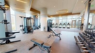 Located at 12008 Inspiration St in Reston, VA, this upscale fitness center spans a generous single floor, offering a comprehensive and thoughtfully designed exercise facility. The space is defined by soaring ceilings, warm wood-paneled accents, and an abundance of natural light streaming through floor-to-ceiling glass windows that frame views of the surrounding Reston landscape.

The main exercise floor is anchored by large, sculptural circular pendant lights that lend a modern, boutique-gym aesthetic. This primary area is equipped with a wide array of strength training machines, cable systems, adjustable benches, power racks, and a full dumbbell rack lining mirrored walls. Rubber flooring in the free weights section ensures safety and durability.

The cardio zone, positioned along the window-lined perimeter, features a row of treadmills, ellipticals, rowing machines (including Concept2 RowERGs), and stair climbers — all with outdoor views to keep workouts engaging. A dedicated functional training area includes medicine balls, kettlebells, and resistance tools.

A separate spin studio provides a distinct space for cycling classes, complete with smart bikes, individual display screens, hardwood-style flooring, and a built-in media wall. Together, these zones create a well-rounded, premium fitness environment suited for all training styles.