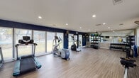This fitness center is a spacious, single-floor facility designed to support a wide range of workout routines. The interior is finished with light wood-look flooring throughout, contributing to a clean, modern aesthetic that complements the functional layout. Recessed lighting illuminates the space evenly, while large windows and glass doors along multiple walls flood the room with natural light and offer views of the surrounding greenery and outdoor areas.

The cardio zone is prominently featured and includes multiple treadmills, elliptical trainers, and a stair climber, all positioned to take advantage of the outdoor-facing windows. A rowing machine and an incline trainer round out the low-impact cardio options. For strength and functional training, the facility provides a multi-station cable machine, a well-stocked dumbbell rack, medicine balls in various weights, resistance accessories, and a dedicated open floor area ideal for stretching or bodyweight exercises.

A large wall mirror on one end enhances the sense of depth in the space, while a wall-mounted television offers entertainment during workouts. The bold navy blue accent walls in portions of the gym add a dynamic visual contrast to the neutral tones. Ceiling fans and ventilation ensure a comfortable environment during intense sessions. The facility is accessible via a glass-paned exterior door, making entry convenient and welcoming.