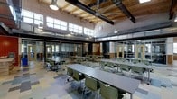 This single-story space is defined by its impressive architectural character and open, flexible layout. The room draws the eye upward to dramatic vaulted ceilings clad in warm natural wood planking, supported by bold exposed steel beams that lend an industrial yet inviting aesthetic. A large ceiling fan anchors the upper space, complementing the industrial-style pendant lighting suspended throughout.

Rows of clerestory windows line the upper walls on multiple sides, flooding the interior with generous natural light while maintaining privacy at ground level. The combination of natural wood, painted masonry walls, and patterned tile flooring creates a layered, durable aesthetic suited for high-traffic use.

The main hall is furnished with long rectangular tables and green stackable chairs arranged in a communal style, accommodating a substantial number of occupants. Along the perimeter, glass-partitioned rooms with wooden doors provide semi-private breakout or auxiliary spaces, visible through transparent walls. Exposed ductwork and mechanical systems are integrated into the ceiling design, reinforcing the space's utilitarian yet thoughtfully designed character. Overall, the space conveys a welcoming, functional atmosphere suited for dining, collaboration, or multipurpose use.
