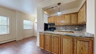 This well-appointed one-bedroom, two-bathroom residence offers a thoughtfully organized single-floor layout with a blend of classic architectural character and functional design. The kitchen serves as a central highlight, featuring warm oak cabinetry with ample upper and lower storage, granite-style countertops, a mosaic tile backsplash, black appliances including a gas range and refrigerator, and a pendant light fixture that adds a polished finishing touch. Rich hardwood floors extend throughout the main living area, complemented by decorative fluted columns that frame the kitchen entry and add a traditional architectural detail. A distinctive solid wood panel door serves as the main entry, adding warmth and character. The spacious primary bedroom is carpeted for comfort and features a classic cast-iron radiator beneath a pair of windows that invite natural light and outdoor views. Two closets provide generous storage, one of which is a walk-in with built-in hanging rods. Both bathrooms include full bathtub and shower combinations with clean white tile surrounds and natural light from windows. The overall ambiance is one of timeless, lived-in character with functional, classic finishes throughout.