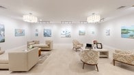 Located at 265 Communication Way in Hyannis, Massachusetts, this single-floor art gallery offers a refined and welcoming environment designed to showcase visual artwork in an elegant, gallery-style setting. The interior is defined by crisp white walls that serve as a pristine backdrop for a rotating collection of colorful paintings, ranging from coastal and nature-inspired works to figurative compositions. Light-toned tile flooring runs throughout the open layout, lending a clean, cohesive aesthetic to the entire space. Illumination is thoughtfully layered, combining sleek adjustable track lighting — ideal for accentuating individual artworks — with statement drum pendant fixtures that add warmth and architectural interest to the ceiling. The gallery is furnished with tastefully curated seating arrangements, including neutral upholstered sofas and armchairs alongside patterned accent chairs, inviting visitors to linger and engage with the art in comfort. A central round coffee table with a decorative base anchors one of the seating areas, adding a touch of warmth and texture. The space flows openly from one seating vignette to the next, creating an intimate yet spacious atmosphere. Private rooms accessible via glass-paneled doors can be seen along one wall, suggesting additional functional areas within the gallery. The overall ambiance is airy, sophisticated, and distinctly curated — a welcoming destination for art enthusiasts on Cape Cod.