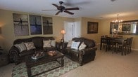 This single-floor residence offers a cohesive and inviting atmosphere defined by warm neutral wall tones and plush carpeted flooring throughout. The living area serves as the heart of the space, anchored by a pair of richly toned dark leather sofas adorned with decorative accent pillows, arranged around a substantial dark wood coffee table. A patterned area rug in muted greens and teals adds a layer of texture and visual interest to the seating area. A ceiling fan with a classic dark finish provides both function and style overhead, complementing the room's earthy, traditional aesthetic. Framed wall art adds character to the living space, contributing to the warm and personal ambiance. Natural light filters into the room, enhancing the welcoming feel of the interior. Just beyond the living area, a glimpse of a dining space is visible, featuring dark bar-height seating that maintains the consistent, rich color palette found throughout. A white interior door leads to adjacent areas of the home, contributing to a clean contrast against the warm-toned walls. Overall, this space presents a traditional, comfortable, and well-coordinated interior ideal for relaxed living.