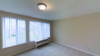 This single-floor unit offers a clean, functional layout with neutral-toned interiors throughout. The main living area features wall-to-wall carpeting in a light beige tone, smooth walls painted in a soft, warm neutral, and standard-height ceilings with flush-mount lighting fixtures. A transition from tile flooring near the entry to carpet in the main living space delineates the layout naturally.

Large windows fitted with vertical blinds allow generous amounts of natural light to fill the rooms, offering views of the surrounding outdoor greenery. Baseboard heating units are positioned beneath the windows, providing efficient climate control across the space. A wall-mounted air conditioning unit in the living area offers supplemental cooling during warmer months.

The walls are clean and unadorned, providing a blank canvas for personalization. White baseboards run consistently throughout, framing the carpeted floors neatly. An electrical panel is visibly mounted on one wall, conveniently accessible. The overall ambiance is modest and practical, with a neutral palette and straightforward finishes that offer a versatile foundation for a variety of interior design preferences.
