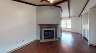 This single-story home showcases a consistent and cohesive design characterized by rich, dark hardwood flooring that runs throughout the entire space, complemented by white walls and warm wood-tone trim and doors. A defining architectural feature is the exposed dark wood beam ceiling detail, present in multiple rooms, lending a rustic yet structured aesthetic to the interior.

The living area is generously proportioned, anchored by a classic wood-burning fireplace with a tiled surround and a decorative wood mantel. Vaulted beam ceilings in this room add vertical dimension, while a ceiling fan provides practical comfort. Natural light enters through a large window, offering views to the outdoors.

Adjacent to the living area, the dining space features its own set of exposed beams and is illuminated by an elegant chandelier. A nearby window further brightens the room.

The galley-style kitchen is well-equipped with ample white cabinetry, gray laminate countertops, a double sink, and white appliances including a refrigerator and range. Crown molding adds a finishing touch.

The bedrooms continue the hardwood flooring and beam ceiling theme, each with closet access. The bathroom includes a wide vanity with gray countertops, wood-framed mirrors, and a separate tub and shower area.