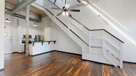 Located at 1450 Greene St in Augusta, Georgia, this single-floor loft-style unit showcases a compelling blend of historic industrial character and contemporary updates. The space is defined by its dramatic soaring ceilings, exposed wooden roof beams, and visible ductwork, all of which reinforce its warehouse-era origins. Richly toned reclaimed hardwood floors extend throughout, adding warmth and texture that contrasts beautifully with the crisp white-painted brick walls.

The open main living area benefits from natural light entering through tall, multi-pane windows, while a ceiling fan provides additional comfort beneath the striking overhead structure. A prominent weathered wood column, its surface layered with aged paint, anchors the space and serves as a striking architectural artifact. Interior staircases with white railings add visual interest and define different levels within the single floor.

The kitchen is outfitted with white cabinetry, dark countertops, and stainless steel appliances including a refrigerator, range, and microwave. A double-basin sink with a bar-height counter provides additional workspace. The bathroom continues the reclaimed hardwood flooring, featuring a white vanity, mirror, and clean modern fixtures, with the same aged column detail present as a design element.