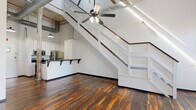 Located at 1450 Greene St in Augusta, Georgia, this single-floor loft-style unit showcases a compelling blend of historic industrial character and contemporary updates. The space is defined by its dramatic soaring ceilings, exposed wooden roof beams, and visible ductwork, all of which reinforce its warehouse-era origins. Richly toned reclaimed hardwood floors extend throughout, adding warmth and texture that contrasts beautifully with the crisp white-painted brick walls.

The open main living area benefits from natural light entering through tall, multi-pane windows, while a ceiling fan provides additional comfort beneath the striking overhead structure. A prominent weathered wood column, its surface layered with aged paint, anchors the space and serves as a striking architectural artifact. Interior staircases with white railings add visual interest and define different levels within the single floor.

The kitchen is outfitted with white cabinetry, dark countertops, and stainless steel appliances including a refrigerator, range, and microwave. A double-basin sink with a bar-height counter provides additional workspace. The bathroom continues the reclaimed hardwood flooring, featuring a white vanity, mirror, and clean modern fixtures, with the same aged column detail present as a design element.