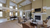 Located at 725 Heather Park Dr in Garner, North Carolina, this single-story clubhouse exudes a refined and welcoming atmosphere from the moment you step inside. The main lounge area is defined by dramatically soaring ceilings with coffered detailing and crisp white crown molding that contrast beautifully against warm golden-tan walls. Tall clerestory windows allow abundant natural light to pour in from multiple directions, creating a bright and airy environment throughout the day. A classic white-framed fireplace serves as a central focal point in the lounge, anchored by a flatscreen television mounted above and flanked by decorative wall sconces that add a warm, ambient glow. The seating area features a curated mix of patterned armchairs, a leather lounge chair, and an ornate area rug, all arranged to encourage conversation and relaxation. A dedicated workspace or business center area is visible to one side, furnished with a dark wood desk and task lighting, offering a functional counterpoint to the lounge's comfort. Large glass-paneled doors lead to additional areas of the building, maintaining a sense of openness and connectivity. Lush potted plants and tasteful décor accents complete the polished, community-centered ambiance of this well-appointed space.