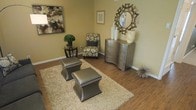 This single-floor space showcases a thoughtfully arranged sitting area that balances comfort and style through a cohesive neutral color palette. The room features warm hardwood flooring throughout, providing a rich foundation that complements the overall design aesthetic.

The walls are finished in a soft, warm beige tone, creating a welcoming backdrop for the carefully selected furnishings. A plush, cream-toned shag area rug anchors the seating arrangement, which includes two metallic leather ottomans with nailhead trim detailing. A patterned accent chair in earthy tones adds visual interest and texture to the space.

A curved metallic chest of drawers serves as a functional yet decorative focal point along one wall, topped with white ceramic vases and a sculptural decor piece. Above it, a starburst-framed mirror adds a bold design element that draws the eye upward. Wall art in abstract and neutral tones complements the room's palette, while a small side table with a potted topiary introduces a natural element. The overall ambiance is warm, polished, and carefully curated, projecting an elegant yet approachable aesthetic.