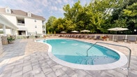 Located at 101 Cool Springs Dr in Elizabethtown, Kentucky, this generously sized outdoor amenity space offers a well-appointed recreational environment designed for both relaxation and social gathering. At the heart of the space is a large, freeform swimming pool with crystal-clear blue water, gentle entry steps, and in-floor features — all surrounded by an expansive, elegantly patterned paver deck that provides ample room for lounging and circulation.

Along the perimeter of the pool deck, a row of cushioned chaise lounges and patio chairs rest beneath multiple market umbrellas, creating shaded retreats framed by mature, lush trees that lend the area a sense of natural privacy. A white fence encloses the pool area, contributing to a clean, resort-like aesthetic.

Adjacent to the pool, a substantial covered pavilion serves as a central gathering hub. The structure features a pitched roof with classic architectural detailing, white-painted wood siding, and prominent columns accented with stacked natural stone bases — adding a refined, craftsman-inspired character. The pavilion provides sheltered seating and dining space, making it ideal for outdoor entertaining in any weather. Discreet utility doors are integrated into the pavilion's design. The surrounding multi-story residential buildings are visible in the backdrop, situating this amenity space as a premier outdoor feature of the broader community.