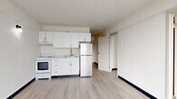 This thoughtfully laid-out one-bedroom, one-bathroom unit offers an efficient and comfortable living experience across a single floor. The open-concept living and kitchen area is anchored by light-toned hardwood flooring and crisp white walls, creating a clean, bright atmosphere throughout. The kitchen is equipped with white cabinetry, a built-in range and oven, a full-size refrigerator, and a sink with ample counter space — all arranged along one wall for a streamlined, functional design.

A central hallway connects the main living area to the private spaces, including the bedroom, bathroom, and closet. The hallway benefits from a large window at its end, drawing in natural light and providing an open, airy feel.

The bedroom is a standout feature of the unit, boasting a floor-to-ceiling corner window that frames expansive outdoor views of the surrounding cityscape and beyond. This generous glazing floods the room with natural light throughout the day. The bedroom also includes dual sliding-door closets, offering well-organized storage space.

The bathroom is finished in a clean, all-white palette and includes a single vanity with cabinet storage, a wall-mounted mirror with overhead lighting, a toilet, and a full bathtub with a glass shower enclosure. Light tile flooring completes the space with a polished, cohesive look.