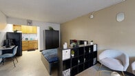 Located along Southwest Fwy in Houston, TX, this 1-bedroom studio offers a well-organized, single-floor layout that makes efficient use of its compact footprint. The open main area is cleverly divided using a black cube shelving unit, visually separating the sleeping zone from the living space without sacrificing openness. The sleeping area features a full-size bed with a tall wardrobe for storage, while the living side includes two modern upholstered accent chairs, a glass-top coffee table, and a floor lamp positioned near a window that brings in natural light. Warm beige walls and a textured ceiling carry throughout the main space, lending a cohesive, neutral tone. The kitchen is tucked to one side and features warm wood cabinetry, granite-style countertops, a cooktop, an under-counter refrigerator, and a stainless steel sink with a ventilation hood above — a practical setup for everyday cooking. The bathroom is clean and bright, finished in white tile with a full tub-shower combination, a grab bar, and a decorative curtain. Three oval wall mirrors in the living area add a modern design touch. A window unit provides climate control throughout the space.