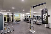Located at 10045 111 St NW in Edmonton, Alberta, this fitness facility offers a thoughtfully organized exercise room across a single floor. The space is finished with durable rubber flooring throughout, bright recessed lighting in the ceiling, and a clean, neutral color palette of white and sage green walls that create an energizing yet focused atmosphere. Full-length mirrors line portions of the walls, visually expanding the room and allowing users to monitor their form during workouts. The facility is comprehensively equipped with a strong selection of Matrix-brand strength training machines, including cable systems, a functional trainer, and a multi-use weight bench. Cardio equipment is well-represented with treadmills, elliptical trainers, and a stationary bike, all positioned to maximize use of the available floor space. A freestanding punching bag and TRX suspension training system add versatility for users seeking varied workout options. A rack of dumbbells provides additional free-weight training capability. The layout is open and efficiently arranged, allowing multiple users to exercise simultaneously without overcrowding. Two access doors and a small window contribute to the functional design of the space, making this fitness room a practical and well-appointed amenity.