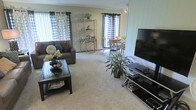 This single-floor home offers a comfortable and functional layout centered around an open-concept living and dining area. The living room is carpeted and furnished with a plush sofa, a glass-top coffee table, and a large flat-screen TV mounted on a light-paneled accent wall, creating a relaxed entertainment space. Decorative touches, including throw pillows, floral arrangements, and a potted indoor plant, add warmth and character to the room. A chrome-frame shelving unit serves as a stylish divider between the living and dining areas, providing both display space and visual separation. The dining area features a bistro-style table with wooden chairs and is bathed in natural light streaming through multiple windows adorned with patterned curtains. The neutral-toned walls and soft carpeting throughout contribute to a cohesive, inviting atmosphere. Track lighting on the ceiling ensures the space is well-lit, complementing the natural light. The overall design balances practicality with homey charm, making it a comfortable everyday living environment.