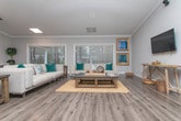 Located at 10300 Springs Ln in Peachtree Corners, Georgia, this single-floor community clubhouse offers a well-appointed gathering space designed for both casual socializing and organized events. The centerpiece of the main area is a dramatic floor-to-ceiling stone fireplace, clad in light-toned stacked stone and framed by an expansive open layout with vaulted ceilings and recessed skylights that flood the space with natural light. Hardwood-style flooring runs throughout, providing a warm, cohesive aesthetic across all areas. The open lounge area features ample room for seating arrangements, complemented by wall-mounted media and decorative framed artwork. An adjacent dining area accommodates a round table with upholstered chairs, positioned alongside windows with views of an outdoor pool area. A long granite-topped bar counter with industrial metal barstools serves as an ideal refreshment station for events or community gatherings. Crown molding and neutral gray walls contribute to a clean, modern-yet-welcoming atmosphere throughout. The space also includes interior doors leading to additional rooms, offering flexibility for various uses. Overall, the clubhouse presents a functional and aesthetically refined environment suited for community activities.