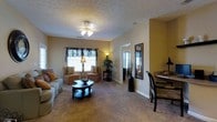 This single-story space features an open and functional layout with warm, neutral tones throughout. The living area is anchored by plush wall-to-wall carpeting in a beige hue, creating a comfortable and cohesive atmosphere. A ceiling fan with an integrated light fixture serves as the central overhead lighting, while a floor lamp near the window provides additional ambient warmth. The room accommodates a sofa, an armchair, and a rounded dark-wood coffee table, forming a welcoming seating arrangement. A window dressed with decorative valances and blinds allows natural light to filter into the space. Textured ceilings add a subtle architectural detail throughout. A tall framed mirror leans against the wall near the hallway, enhancing the sense of depth and openness. The layout transitions naturally toward what appears to be additional rooms, visible through a bright doorway in the background. To the right, a dining area with a small table and chairs is partially visible, suggesting a seamless connection between living and dining spaces. Decorative accents, including potted plants and floor vases, contribute to the overall warmth of the interior.