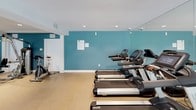 This fitness center is designed to support a comprehensive workout experience within a single-level layout. The space is divided into distinct functional zones, with one area dedicated to cardiovascular equipment and another to strength training machines. The cardio section features multiple treadmills and stationary bikes arranged in an orderly row, while the strength training area houses a variety of cable and resistance machines along with a weight bench and free weights.

Bold teal accent walls provide a visually energizing atmosphere, contrasting with white trim and a neutral-toned floor that runs throughout the space. Recessed lighting is evenly distributed across the white ceiling, ensuring consistent, well-lit conditions for exercise. Full-length mirrors line one wall, enhancing the sense of openness and allowing users to monitor their form during workouts.

A standout feature is the large sliding glass doors and floor-to-ceiling windows at one end of the space, which flood the area with natural light and offer views of an adjacent outdoor terrace. The overall ambiance is clean, modern, and functional, reflecting a thoughtfully designed environment suited for a variety of fitness routines.