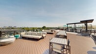 The Trove Sun Deck in Arlington, VA is an expansive rooftop amenity space designed to maximize outdoor living and entertainment. The deck features warm composite wood decking throughout, complemented by a sleek, modern architectural aesthetic with stone cladding, floor-to-ceiling glass facades, and clean contemporary lines.

Multiple distinct seating zones are thoughtfully arranged across the space. Plush, neutral-toned sectional sofas and lounge chairs provide relaxed gathering areas, while long communal dining tables with ample seating accommodate larger groups. A covered pergola-style outdoor kitchen and grilling station offers a shaded hub for cooking and socializing.

A dedicated pool area features lounge chairs and teal umbrellas, separated from the main deck by a transparent glass railing that preserves unobstructed views. Adjacent to the building's glass entry, a cozy seating nook with woven accent chairs surrounds an outdoor fireplace set into a stone feature wall, creating an intimate retreat.

The deck offers panoramic views of the surrounding Arlington neighborhood, lush tree canopy, and the broader Washington, D.C. metro skyline in the distance. Open skies and abundant natural light define the atmosphere throughout the day, making this rooftop an ideal venue for both casual relaxation and social gatherings.