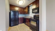 This single-floor unit showcases a cohesive design defined by polished terrazzo flooring that runs seamlessly throughout the space, lending a clean, durable foundation to every room. The kitchen is efficiently laid out in an L-shaped configuration, equipped with rich dark-stained wood cabinetry extending to the ceiling for generous storage. Black appliances — including a refrigerator, electric range, and built-in microwave — complement the granite-style countertops, while floating glass shelves add a contemporary accent. A flush-mount ceiling light and light blue backsplash complete the functional yet stylish space. The bedroom is notably spacious, featuring elevated ceilings, a ceiling fan, and a row of large windows that frame striking urban skyline views and flood the room with natural light. A built-in closet provides ample storage. The bathroom includes a full tub-shower combination, a dark-wood vanity with an integrated sink, and a large mirror. A dedicated in-unit laundry closet accommodates a stacked front-loading washer, offering added convenience. The overall layout, visible in the 3D model, is well-organized and efficiently utilizes the available footprint.
