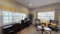 This single-floor office space is designed to accommodate both staff operations and guest interactions in a polished, professional environment. The layout includes a glass-walled interior office that provides visual transparency while maintaining a defined workspace, allowing staff to remain connected to the broader common areas. Adjacent to the glass partition, an open area with large tile flooring leads into a comfortable lounge and breakroom space, visible through the open-plan design.

The primary office and lounge area features warm wood-look flooring and is bathed in natural light from multiple large windows dressed with layered Roman shades and grommet curtains in a coordinated gray, white, and yellow palette. A dedicated workstation accommodates dual monitors and office equipment along a dark wood credenza, paired with ergonomic seating. The lounge area is furnished with a pair of gray leather wingback chairs accented with yellow throw pillows, centered around a glass-top oval coffee table — creating an inviting space for consultations or casual meetings.

Recessed lighting throughout complements the abundant natural light, while tall vaulted ceilings in the main office area add a sense of openness. A glass-paned exterior door provides direct outdoor access and additional natural light. The space maintains a consistent, well-curated aesthetic throughout.