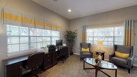 This single-floor office space is designed to accommodate both staff operations and guest interactions in a polished, professional environment. The layout includes a glass-walled interior office that provides visual transparency while maintaining a defined workspace, allowing staff to remain connected to the broader common areas. Adjacent to the glass partition, an open area with large tile flooring leads into a comfortable lounge and breakroom space, visible through the open-plan design.

The primary office and lounge area features warm wood-look flooring and is bathed in natural light from multiple large windows dressed with layered Roman shades and grommet curtains in a coordinated gray, white, and yellow palette. A dedicated workstation accommodates dual monitors and office equipment along a dark wood credenza, paired with ergonomic seating. The lounge area is furnished with a pair of gray leather wingback chairs accented with yellow throw pillows, centered around a glass-top oval coffee table — creating an inviting space for consultations or casual meetings.

Recessed lighting throughout complements the abundant natural light, while tall vaulted ceilings in the main office area add a sense of openness. A glass-paned exterior door provides direct outdoor access and additional natural light. The space maintains a consistent, well-curated aesthetic throughout.