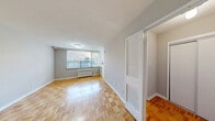 Located at 145 Jameson Ave in Toronto's vibrant Parkdale neighbourhood, this single-floor unit offers a practical and efficient layout comprising a living room, bathroom, and hallway. The living room is the heart of the space, featuring warm parquet wood flooring laid in a classic block pattern that extends throughout. Large vertical-blind-dressed windows invite natural light and offer street-level views of the surrounding neighbourhood, while a radiator beneath ensures comfortable heating. Neutral grey walls provide a clean, versatile backdrop throughout the unit. The hallway connects the main living area to the bathroom and includes a louvred bifold closet door, offering convenient storage. A solid wood entry door with hardware anchors the entrance. The bathroom is neatly appointed with a tiled tub-shower combination, a vanity featuring a granite-look countertop with an integrated sink, and tiled walls in neutral tones, complemented by a mirrored medicine cabinet. The overall ambiance is understated and functional, with a consistent colour palette of warm wood tones and soft grey that lends the space a calm, cohesive feel throughout.