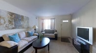 This single-floor space presents a thoughtfully arranged living room that serves as the central gathering area. The room is finished with wall-to-wall carpeting in a neutral beige tone, contributing to a warm and comfortable atmosphere throughout. Soft, light-colored walls provide a clean backdrop that enhances the sense of openness in the space.

The seating area is anchored by a large, cushioned sofa adorned with blue accent pillows, paired with a matching loveseat, both positioned around a dark-toned oval coffee table that adds a subtle contrast to the lighter surroundings. A decorative floral arrangement on the coffee table adds a natural, inviting touch. An abstract canvas artwork on the wall introduces visual interest and a hint of color to the space.

Natural light filters into the room through a window dressed with sheer and decorative curtains, positioned adjacent to a paneled front entry door featuring a glass transom detail. A classic table lamp provides supplemental ambient lighting. The layout is efficient and practical, with clear sightlines across the room, making the space feel open and easy to navigate.