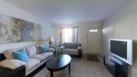 This single-floor space presents a thoughtfully arranged living room that serves as the central gathering area. The room is finished with wall-to-wall carpeting in a neutral beige tone, contributing to a warm and comfortable atmosphere throughout. Soft, light-colored walls provide a clean backdrop that enhances the sense of openness in the space.

The seating area is anchored by a large, cushioned sofa adorned with blue accent pillows, paired with a matching loveseat, both positioned around a dark-toned oval coffee table that adds a subtle contrast to the lighter surroundings. A decorative floral arrangement on the coffee table adds a natural, inviting touch. An abstract canvas artwork on the wall introduces visual interest and a hint of color to the space.

Natural light filters into the room through a window dressed with sheer and decorative curtains, positioned adjacent to a paneled front entry door featuring a glass transom detail. A classic table lamp provides supplemental ambient lighting. The layout is efficient and practical, with clear sightlines across the room, making the space feel open and easy to navigate.