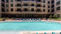 This amenity space features two distinct areas designed to support both relaxation and active living. The outdoor pool area presents a generously sized, crystal-clear swimming pool with a depth of 4'6", bordered by a spacious sun deck lined with teal and navy lounge chairs. The pool is set against the backdrop of a modern multi-story building featuring stacked balconies, floor-to-ceiling glass storefronts at ground level, and contemporary masonry façades — creating a resort-like atmosphere ideal for outdoor enjoyment.

Adjacent to the pool area, the fitness center is well-appointed and spans a comfortable open layout. The space is brightened by multiple large windows fitted with white blinds, allowing natural light to flow in while offering views of the outdoors. Ceiling fans provide additional airflow throughout. The fitness center is equipped with a comprehensive selection of strength training machines, a multi-functional cable and power rack system, a full dumbbell rack, medicine balls, battle ropes, stability balls, and a Precor stationary bike. A dedicated cardio area houses multiple treadmills paired with a wall-mounted television. Floor-to-ceiling mirrors visually expand the space, and patterned carpet tile flooring adds a modern, polished finish throughout.