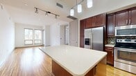 This 990-square-foot, single-floor apartment at Griffis Uptown on Carlisle Street in Dallas, TX offers 3 bedrooms and 2 bathrooms with a well-designed layout. The combined kitchen and living room features warm hardwood flooring throughout, while the kitchen is equipped with rich dark cabinetry, stainless steel appliances including a refrigerator, range, and over-the-range microwave, complemented by a mosaic tile backsplash and light countertops. The living area benefits from natural light and access to an outdoor balcony through a glass door. The primary bedroom spans 160 square feet and connects to a full bathroom finished with large-format tile flooring, a soaking tub with stone-tile surround, and a dark wood vanity with a single sink. A second full bathroom mirrors this design. Bedrooms feature plush carpet flooring and ceiling fans, with multiple windows offering views of the surrounding Dallas skyline. Two generously sized closets with wire shelving systems provide substantial storage. A hallway connects the rooms, completing this functional and stylish urban apartment.