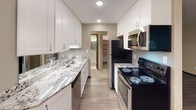 This single-floor residential unit offers a thoughtfully updated interior with a cohesive design throughout. The galley-style kitchen is a standout feature, boasting granite countertops, white shaker cabinetry, and stainless steel appliances including a range, microwave, and dishwasher. A double basin sink and recessed lighting add to the kitchen's functional appeal, while a hallway leads directly to a bathroom and a spacious walk-in closet or storage area beyond.

The bathroom is well-appointed with a granite-topped vanity, white cabinetry, a full tub and shower combo with tile surround, and a wide mirror illuminated by a multi-bulb vanity light fixture. The warm, neutral tones create a clean and inviting atmosphere.

The living areas include a bright room with wide windows that frame outdoor greenery, fitted with carpet flooring and baseboard heating. An additional room features dark hardwood-style plank flooring, a textured ceiling, and louvered bifold closet doors, providing generous storage. The main living area is open and airy, with multiple windows along one wall allowing natural light to fill the space, complemented by hardwood-style flooring and a wall-mounted air conditioning unit. Together, the rooms provide a comfortable, move-in ready environment.