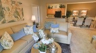 This single-floor residence offers a thoughtfully designed open-concept layout that connects the living, dining, and kitchen areas into one cohesive space. The living room serves as the central gathering area, featuring plush neutral-toned sofas accented with soft blue and floral decorative pillows, layered over a richly patterned area rug. A round gold coffee table anchors the seating arrangement, adding an elegant touch to the relaxed atmosphere. Natural light enhances the warm beige walls, creating an inviting and comfortable ambiance throughout. A pass-through counter connects the living area to the kitchen, which is outfitted with honey-toned wood cabinetry, dark appliances, and ample counter space, making it both functional and visually appealing. Decorative accents above the cabinetry add personality to the space. Adjacent to the living room, a glimpse of the dining area reveals a light-toned table with upholstered seating, ideal for casual meals. The overall design balances practicality with a welcoming aesthetic, making this a comfortable and well-appointed single-level home.